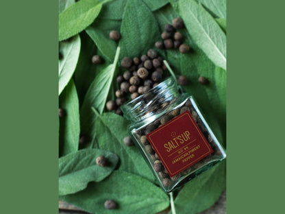 Glass jar of Salt's Up No 90 Jamaican piment pepper on fresh green leaves with scattered peppercorns