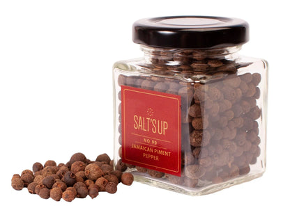 Glass jar of Salt's Up Jamaican Piment Pepper with a pile of loose brown peppercorns on white background