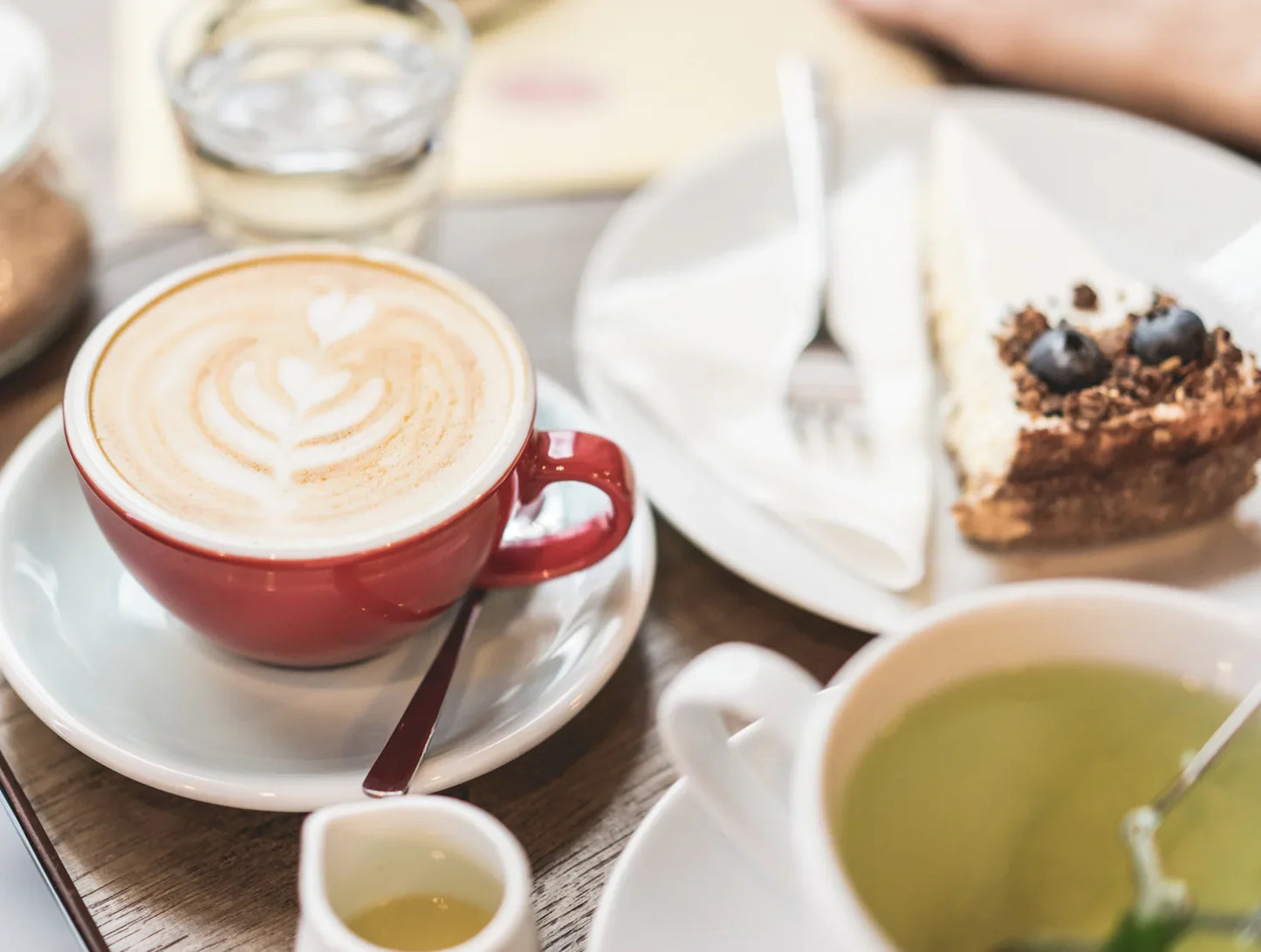 Close-up of latte art in red cup, slice of blueberry cheesecake, and cup of green tea on wooden table