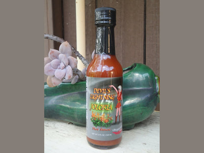 Devil's Lightning Jolokia hot sauce bottle on a rustic shelf with succulent plant and green ceramic.